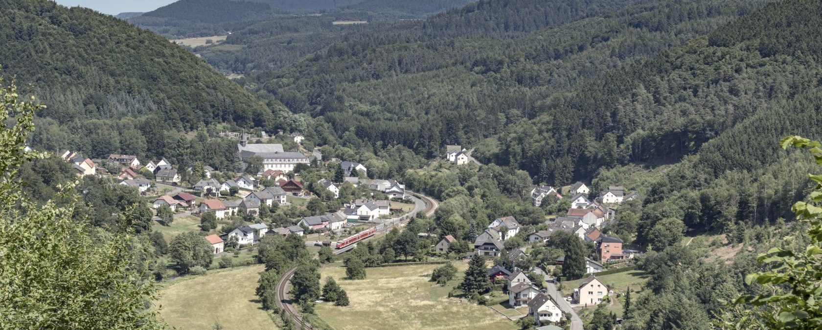 Vue panoramique sur un village situé dans une vallée verdoyante, entouré de collines boisées et de prairies. Un train traverse le village., © Rudolf Höser Vue panoramique sur un village situé dans une vallée verdoyante, entouré de collines boisées et de prairies. Un train traverse le village., © Rudolf Höser