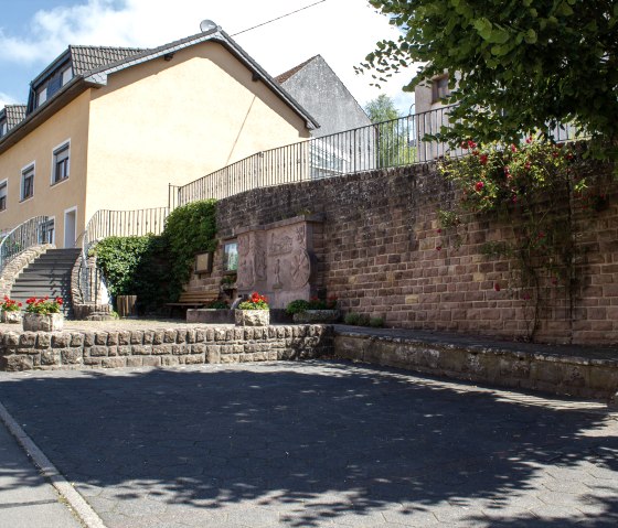 Un parking vide à côté d'une église. On peut voir un bâtiment jaune avec un escalier et un mur de pierre avec des fleurs et des arbres., © TI Bitburger Land - M.Mayer Un parking vide à côté d'une église. On peut voir un bâtiment jaune avec un escalier et un mur de pierre avec des fleurs et des arbres., © TI Bitburger Land - M.Mayer