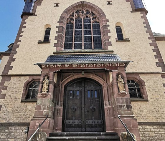 Eingang der Pfarrkirche Maria Königin in Dudeldorf mit steinerner Treppe, verzierten Holztüren und Statuen. Gotische Fenster und Ziegelsteindetails., © Melanie Lonien Eingang der Pfarrkirche Maria Königin in Dudeldorf mit steinerner Treppe, verzierten Holztüren und Statuen. Gotische Fenster und Ziegelsteindetails., © Melanie Lonien