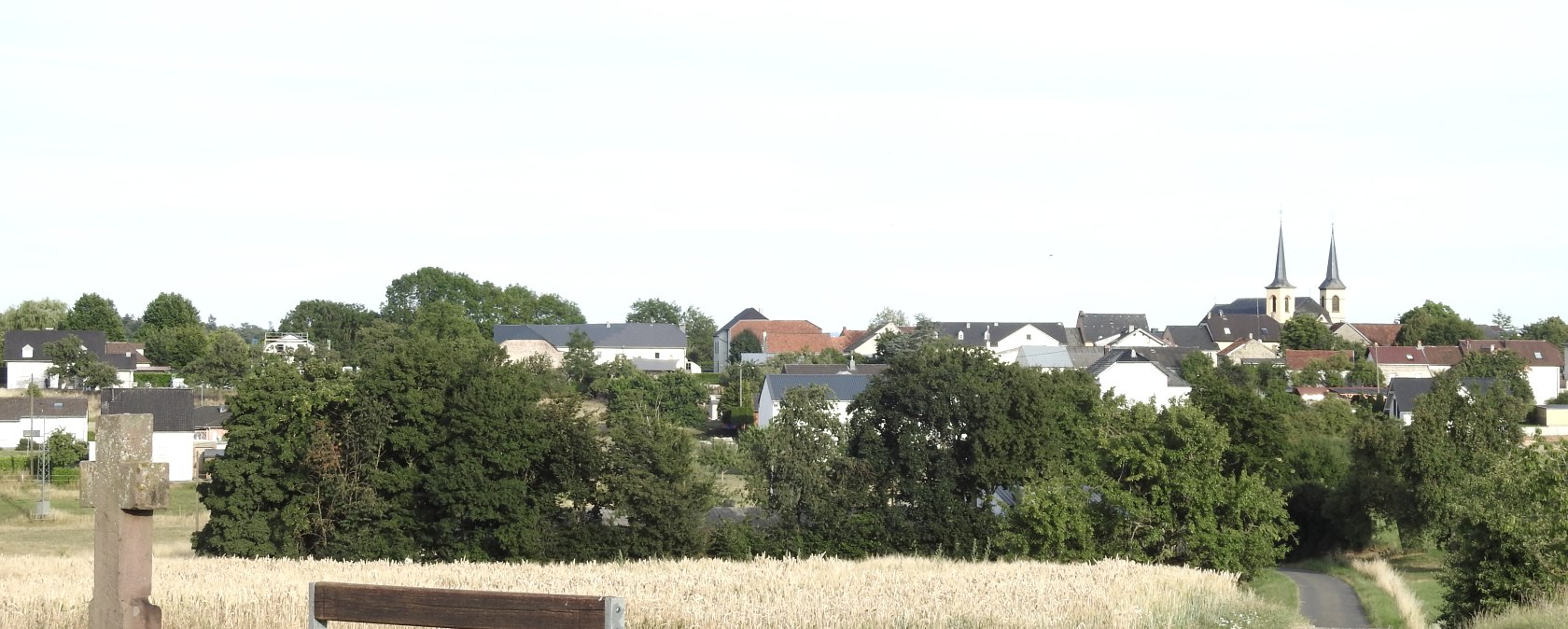 Een bank en een kruis staan voor een veld. Op de achtergrond is een dorp te zien met een kerk en twee torens., © Thomas Neises Een bank en een kruis staan voor een veld. Op de achtergrond is een dorp te zien met een kerk en twee torens., © Thomas Neises