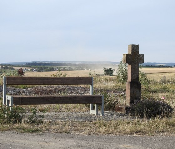 Een houten bank en een stenen kruis staan op een zandpad. Op de achtergrond is een maaidorser te zien in een veld., © Thomas Neises Een houten bank en een stenen kruis staan op een zandpad. Op de achtergrond is een maaidorser te zien in een veld., © Thomas Neises