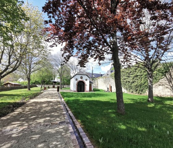 Ein gepflegter Park mit Bäumen, grünem Rasen und einem kleinen weißen Gebäude. Ein gepflasterter Weg führt durch den Park., © Melanie Lonien Ein gepflegter Park mit Bäumen, grünem Rasen und einem kleinen weißen Gebäude. Ein gepflasterter Weg führt durch den Park., © Melanie Lonien