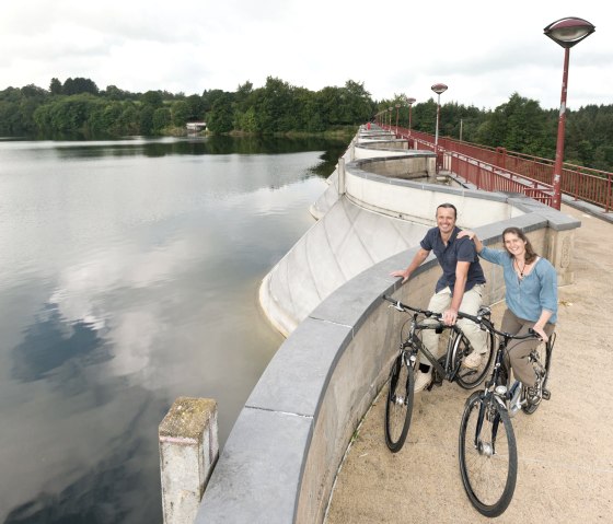 Zwei Personen mit Fahrrädern stehen auf der Staumauer des Stausees Bütgenbach. Im Hintergrund sind Wasser und Bäume zu sehen., © vennbahn.eu Zwei Personen mit Fahrrädern stehen auf der Staumauer des Stausees Bütgenbach. Im Hintergrund sind Wasser und Bäume zu sehen., © vennbahn.eu
