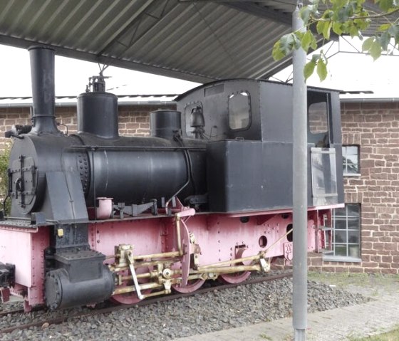 Een oude stoomlocomotief staat onder een afdak voor een bakstenen gebouw. De locomotief is zwart en roze geschilderd., © Eifelverein Ortsgruppe Speicher Een oude stoomlocomotief staat onder een afdak voor een bakstenen gebouw. De locomotief is zwart en roze geschilderd., © Eifelverein Ortsgruppe Speicher