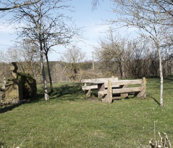 Rastplatz mit Holzbänken und steinernem Kreuz auf einer Wiese, umgeben von kahlen Bäumen unter blauem Himmel., © M. Bach Rastplatz mit Holzbänken und steinernem Kreuz auf einer Wiese, umgeben von kahlen Bäumen unter blauem Himmel., © M. Bach