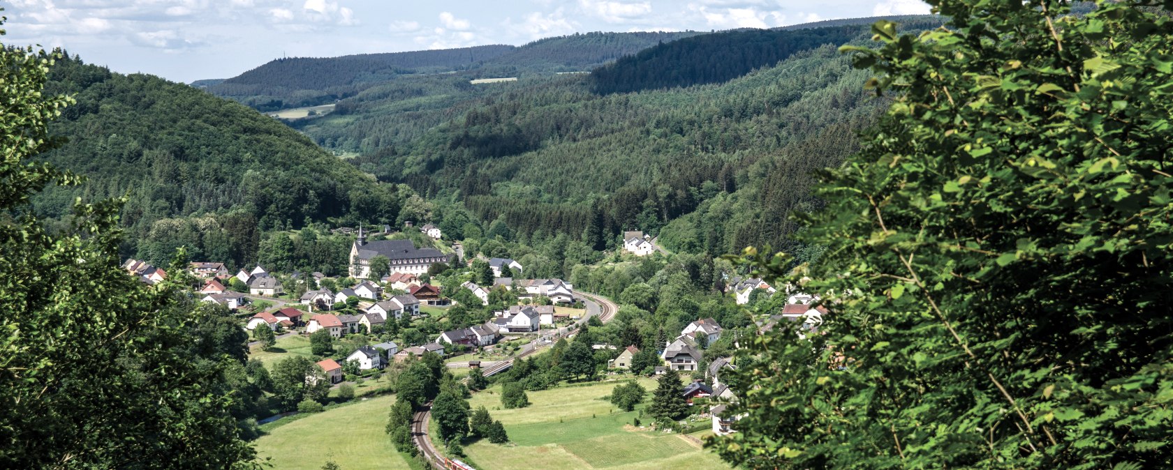 Panoramablick auf ein Dorf inmitten grüner Hügel und Wälder. Ein Zug fährt auf einer Bahnstrecke durch die Landschaft., © TI Bitburger Land Panoramablick auf ein Dorf inmitten grüner Hügel und Wälder. Ein Zug fährt auf einer Bahnstrecke durch die Landschaft., © TI Bitburger Land