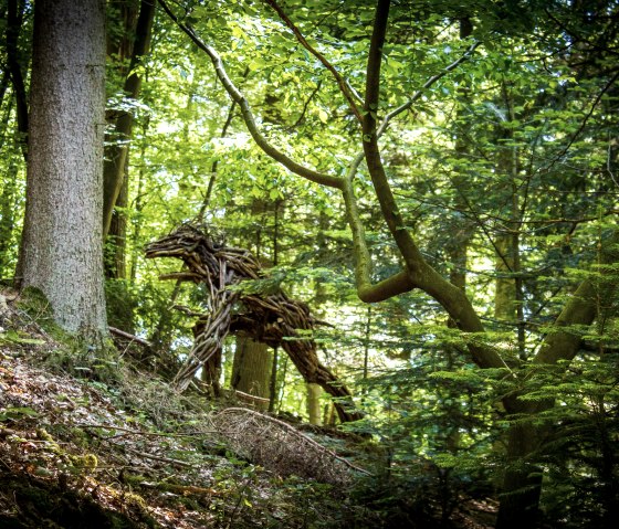 Een houten sculptuur in het bos dat lijkt op een dier, omringd door dicht gebladerte en bomen, onderdeel van de Kyllburg kunstroute., © TI Bitburger Land_M.Mayer Een houten sculptuur in het bos dat lijkt op een dier, omringd door dicht gebladerte en bomen, onderdeel van de Kyllburg kunstroute., © TI Bitburger Land_M.Mayer