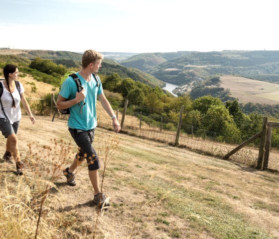 Zwei Personen wandern auf einem Hügel mit Blick auf eine Flusslandschaft. Die Umgebung ist grün und hügelig, der Himmel ist klar., © Eifel Tourismus GmbH, D. Ketz Zwei Personen wandern auf einem Hügel mit Blick auf eine Flusslandschaft. Die Umgebung ist grün und hügelig, der Himmel ist klar., © Eifel Tourismus GmbH, D. Ketz