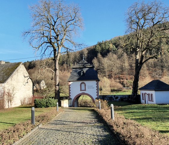 Porte d'entrée du monastère Saint-Thomas avec chemin pavé, entourée d'arbres et de bâtiments, devant une colline boisée., © TI BItburger Land - Steffi Wagner Porte d'entrée du monastère Saint-Thomas avec chemin pavé, entourée d'arbres et de bâtiments, devant une colline boisée., © TI BItburger Land - Steffi Wagner