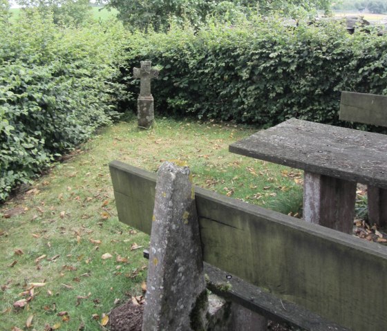 Een stenen kruis staat op een weiland naast een verweerde houten tafel, omringd door dichte heggen en bomen., © M. Bach Een stenen kruis staat op een weiland naast een verweerde houten tafel, omringd door dichte heggen en bomen., © M. Bach