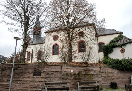 Die Kirche in Wißmannsdorf mit roten Fensterrahmen, umgeben von kahlen Bäumen und einer Steinmauer im Vordergrund., © TI Bitburger Land Die Kirche in Wißmannsdorf mit roten Fensterrahmen, umgeben von kahlen Bäumen und einer Steinmauer im Vordergrund., © TI Bitburger Land
