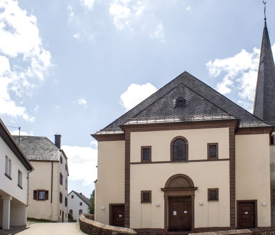 Die Pfarrkirche St. Peter in Neidenbach mit spitzem Turm, umgeben von Wohnhäusern, unter einem blauen Himmel mit Wolken., © TI Bitburger Land, M.Mayer Die Pfarrkirche St. Peter in Neidenbach mit spitzem Turm, umgeben von Wohnhäusern, unter einem blauen Himmel mit Wolken., © TI Bitburger Land, M.Mayer