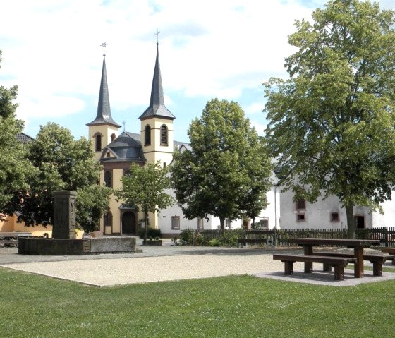 Ein idyllischer Dorfplatz in Idenheim mit einer Kirche im Hintergrund, umgeben von Bäumen. Im Vordergrund sind Bänke und ein Brunnen zu sehen., © Ingrid Penning Ein idyllischer Dorfplatz in Idenheim mit einer Kirche im Hintergrund, umgeben von Bäumen. Im Vordergrund sind Bänke und ein Brunnen zu sehen., © Ingrid Penning