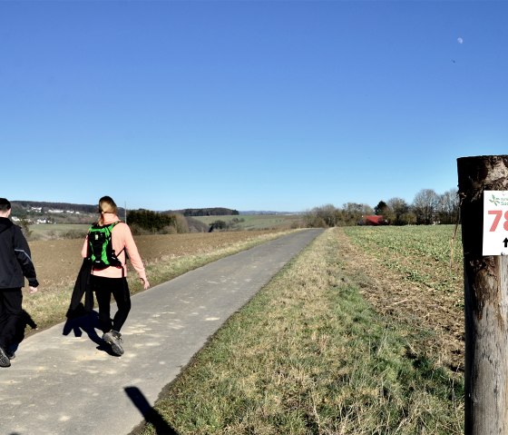 Twee wandelaars op een pad in het natuurpark Zuid-Eifel. Een paal met het nummer 78 staat aan de rand van het pad. De lucht is helder en blauw., © TI Bitburger Land Twee wandelaars op een pad in het natuurpark Zuid-Eifel. Een paal met het nummer 78 staat aan de rand van het pad. De lucht is helder en blauw., © TI Bitburger Land