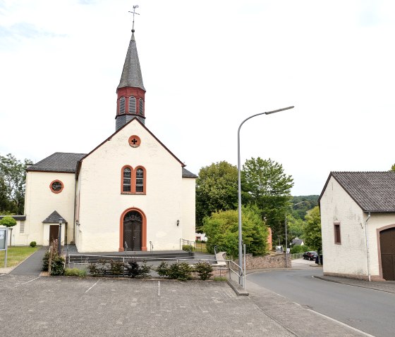 Een kerk in Wißmannsdorf met een spitse toren, omringd door bomen. Ervoor een lege parkeerplaats en een weg. Een ander gebouw aan de rechterkant., © TI Bitburger Land Een kerk in Wißmannsdorf met een spitse toren, omringd door bomen. Ervoor een lege parkeerplaats en een weg. Een ander gebouw aan de rechterkant., © TI Bitburger Land