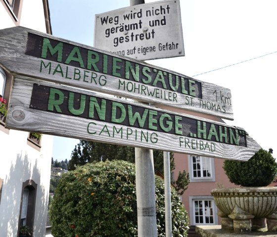 Een houten wegwijzer in Kyllburg wijst de weg naar de Marien-Säule en de Rundwege Hahn. Op de achtergrond zijn gebouwen en planten te zien., © TI Bitburger Land Een houten wegwijzer in Kyllburg wijst de weg naar de Marien-Säule en de Rundwege Hahn. Op de achtergrond zijn gebouwen en planten te zien., © TI Bitburger Land