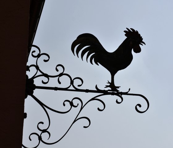 Silhouet van een haan op een artistiek versierd metalen bord voor een heldere hemel., © TI Bitburger Land Silhouet van een haan op een artistiek versierd metalen bord voor een heldere hemel., © TI Bitburger Land