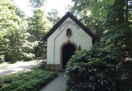 Eine kleine Kapelle aus dem Jahr 1877 steht im Wald, umgeben von Bäumen und einem Weg. Die Sonne scheint durch das Blätterdach. Eine kleine Kapelle aus dem Jahr 1877 steht im Wald, umgeben von Bäumen und einem Weg. Die Sonne scheint durch das Blätterdach.