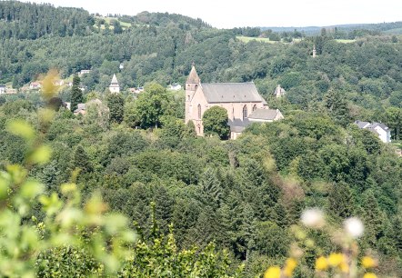 Blick auf eine Kirche, umgeben von dichten Wäldern und Hügeln, unter blauem Himmel. Im Vordergrund sind unscharfe Pflanzen zu sehen., © Tourist-Info Bitburger Land M.Mayer Blick auf eine Kirche, umgeben von dichten Wäldern und Hügeln, unter blauem Himmel. Im Vordergrund sind unscharfe Pflanzen zu sehen., © Tourist-Info Bitburger Land M.Mayer