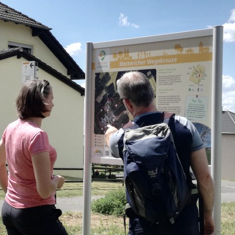 Deux randonneurs se tiennent devant un panneau de randonnée portant l'inscription 'Mettericher Wegekreuze' et s'informent sur l'itinéraire. Des maisons sont visibles à l'arrière-plan., © Tourist-Information Bitburger Land Deux randonneurs se tiennent devant un panneau de randonnée portant l'inscription 'Mettericher Wegekreuze' et s'informent sur l'itinéraire. Des maisons sont visibles à l'arrière-plan., © Tourist-Information Bitburger Land