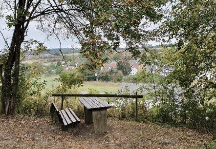 Un banc en bois sous un arbre offre une vue sur le village de Gransdorf dans un paysage verdoyant et vallonné., © TI BItburger Land - Steffi Wagner Un banc en bois sous un arbre offre une vue sur le village de Gransdorf dans un paysage verdoyant et vallonné., © TI BItburger Land - Steffi Wagner