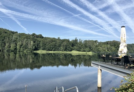 Le lac de barrage de Bitburg aux eaux calmes, entouré de forêt. Une terrasse avec des tables et un parasol se dresse dans l'image. Ciel dégagé avec des traînées de condensation., © TI Bitburger Land Le lac de barrage de Bitburg aux eaux calmes, entouré de forêt. Une terrasse avec des tables et un parasol se dresse dans l'image. Ciel dégagé avec des traînées de condensation., © TI Bitburger Land