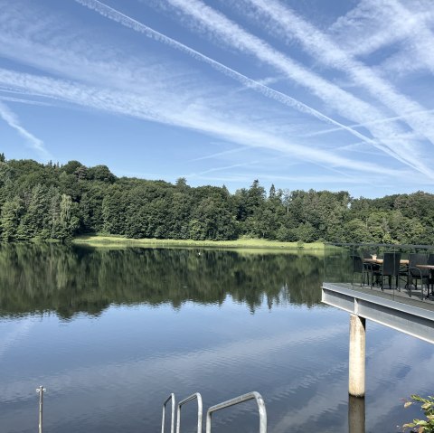 Le lac de barrage de Bitburg aux eaux calmes, entouré de forêt. Une terrasse avec des tables et un parasol se dresse dans l'image. Ciel dégagé avec des traînées de condensation., © TI Bitburger Land Le lac de barrage de Bitburg aux eaux calmes, entouré de forêt. Une terrasse avec des tables et un parasol se dresse dans l'image. Ciel dégagé avec des traînées de condensation., © TI Bitburger Land