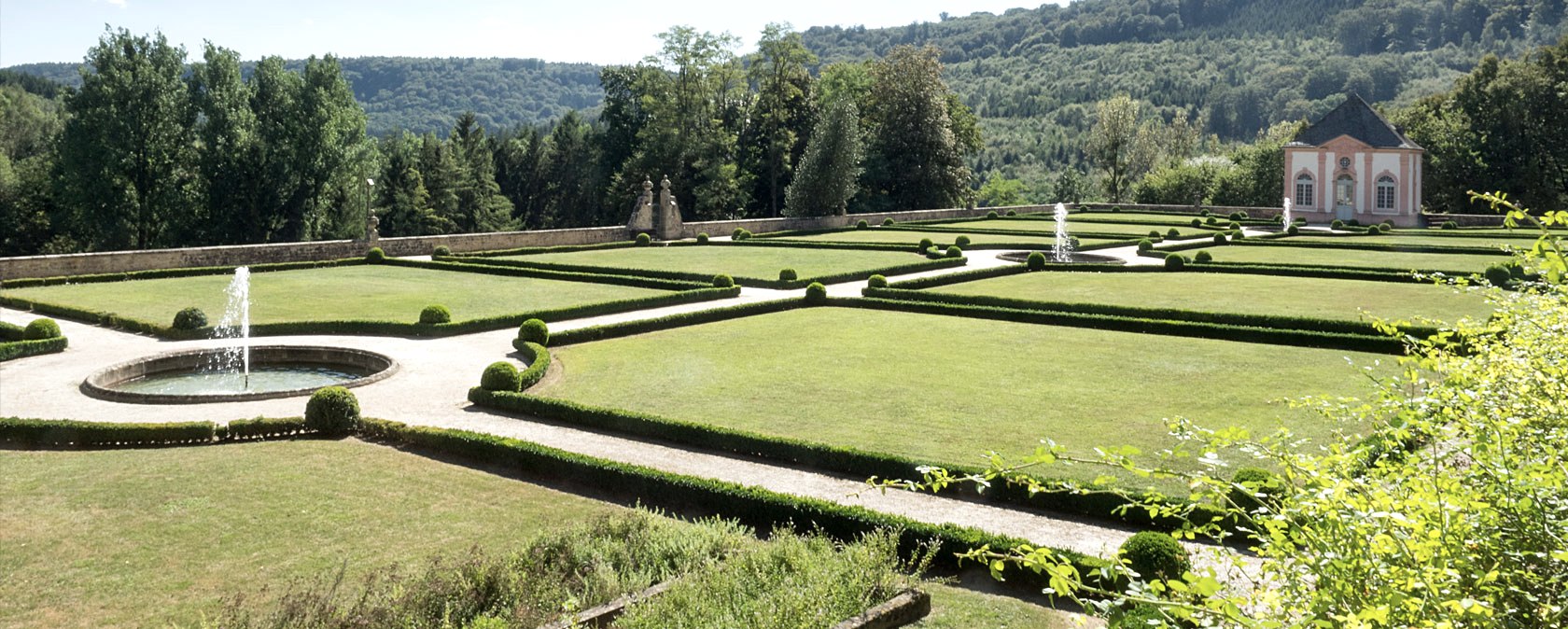 Een goed onderhouden Franse tuin met symmetrische hagen, fonteinen en een paviljoen op de achtergrond, omringd door bos., © V. Teuschler Een goed onderhouden Franse tuin met symmetrische hagen, fonteinen en een paviljoen op de achtergrond, omringd door bos., © V. Teuschler