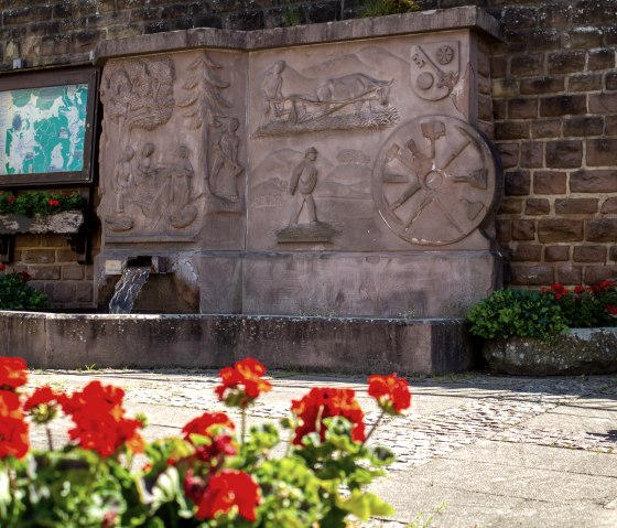 Ein Steinbrunnen mit kunstvollen Reliefs und flie&szlig;endem Wasser. Rote Blumen im Vordergrund, eine Infotafel und eine Bank daneben., &copy; TI Bitburger Land - Monika Mayer