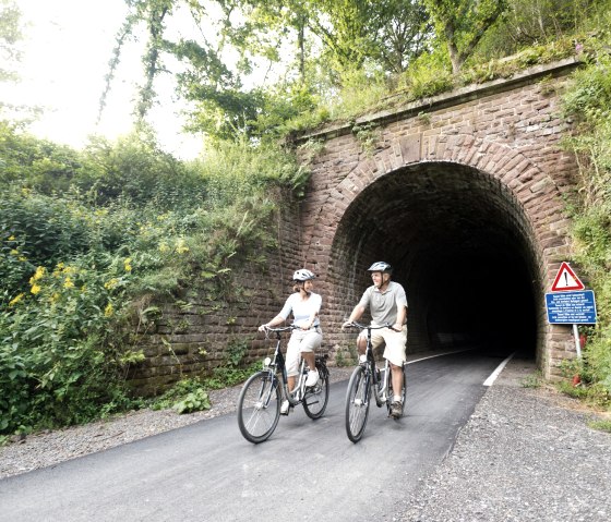 Deux cyclistes sortent d'un tunnel couvert de v&eacute;g&eacute;tation sur la Vennbahn pr&egrave;s de Lommersweiler. Un panneau d'avertissement se trouve sur le bord., &copy; vennbahn.eu