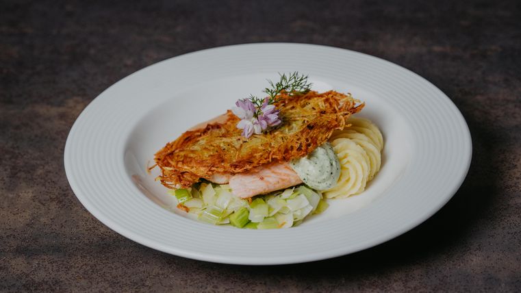 A beautiful plate with fried salmon, surrounded by puree and leeks. The dish is decorated with herbs and an edible flower.