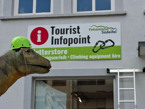 Ein Tourist-Infopunkt mit einem Schild, das Kletterausrüstung anbietet. Im Vordergrund steht eine große Dinosaurierfigur mit einem grünen Helm.