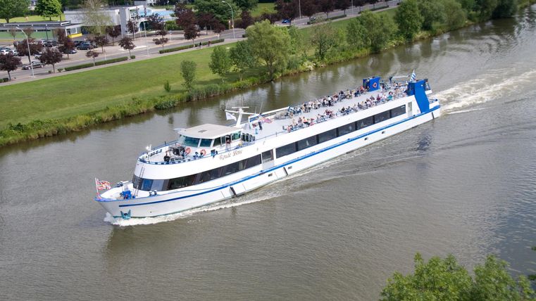 Een grote passagiersboot vaart op een rustige rivier. Op de achtergrond zijn groene bomen en een pad te zien.