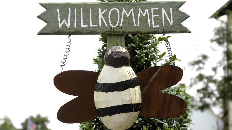 A sign with the inscription "WELCOME" and a wooden figure of a bee. The bee has black and white stripes and outward-facing wings.