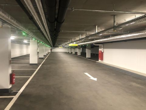 Ein leerer Parkplatz mit mehreren Parkplätzen und einer Decke mit Rohren. Die Beleuchtung ist modern und es gibt grüne und gelbe Lichtsignale.