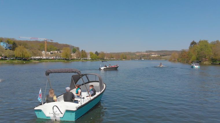 Een boot vaart op een rustige rivier onder een heldere blauwe hemel. Op de achtergrond zijn enkele bomen en kleine heuvels te zien.