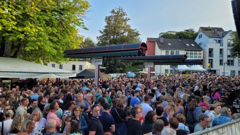 A large crowd gathers at a venue. In the background, some buildings and trees are visible.