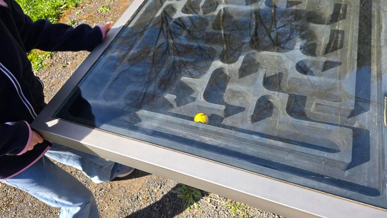 A person is holding a large labyrinthine game board with a small yellow ball. The ground is green and the weather seems to be sunny.