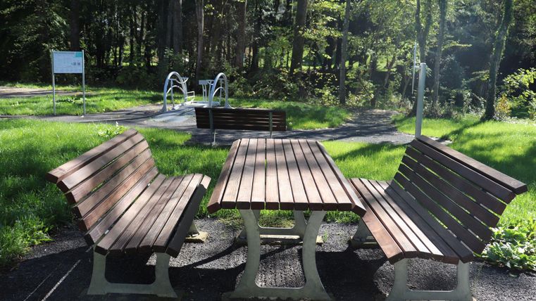 Ein gepflasterter Sitzbereich mit Holzbänken und einem Tisch im Wald. Im Hintergrund sind Grünflächen und Spielgeräte zu sehen.