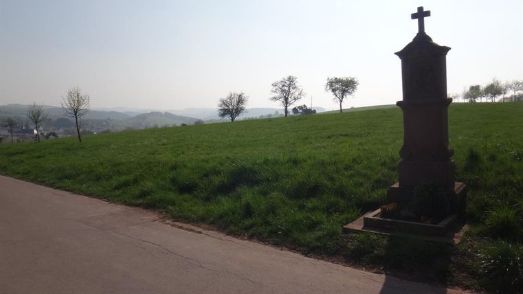 Ein friedlicher Feldweg mit einer Statue und Bäumen im Hintergrund. Der Himmel ist klar und die Landschaft wirkt ruhig.