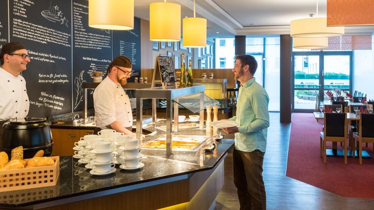 Een moderne eetruimte met een buffet. Een gast staat bij het buffet en wordt door twee koks bediend.