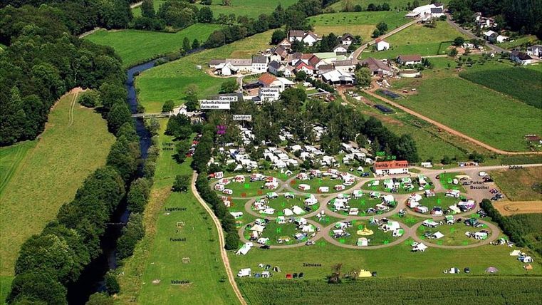 An aerial view of a campsite with many tents and RVs in a green landscape. In the background, a small river flows and there are some buildings visible.