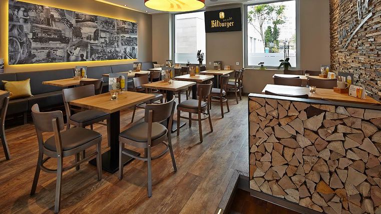 Een modern restaurant met een houten vloer en gezellige tafels. Aan de muur hangt een grote foto en er is een decoratieve houtstapel te zien.