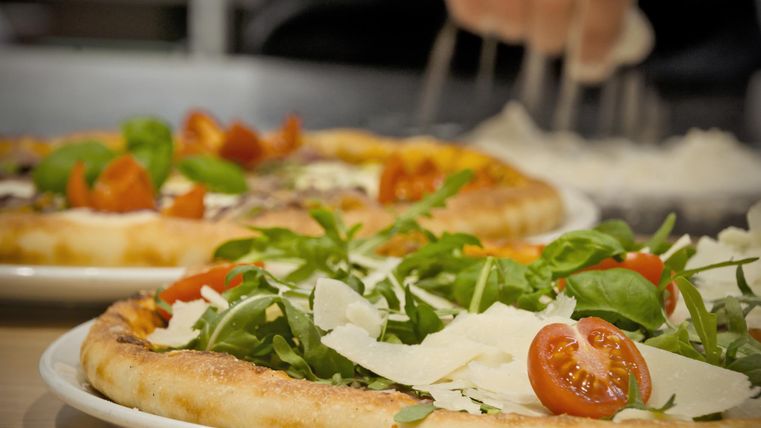 Two delicious pizzas with fresh vegetables and arugula on light plates. In the background, pizza dough is being prepared.