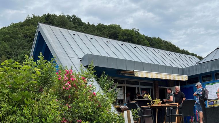 Een modern gebouw met een schuin dak in een natuurlijke omgeving. Mensen staan buiten en genieten van de sfeer.