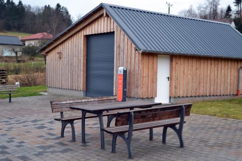Een houten gebouw met een grote rolluik en een witte poort. Voor het gebouw staan twee houten tafels en banken op een betegeld gebied.