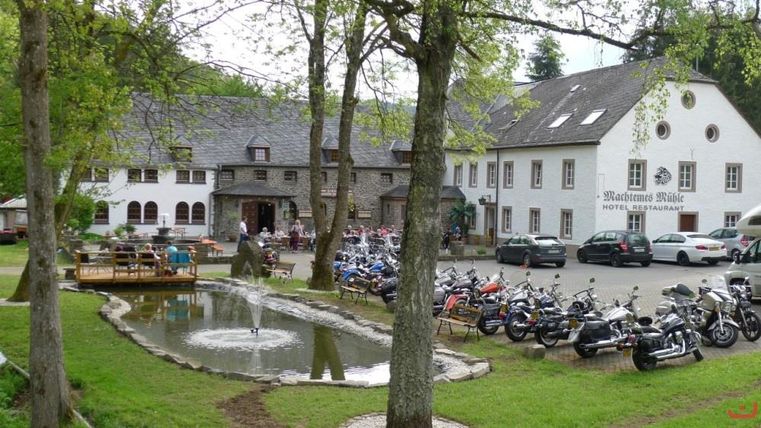 A picturesque restaurant with a small pond and trees. Many motorcycles are parked in front of the building.