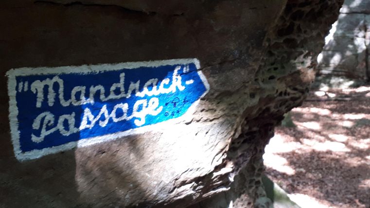 Un panneau bleu avec l'inscription "Mandrasch-Passage" indique le chemin. Il se trouve sur une surface rocheuse dans une zone forestière.