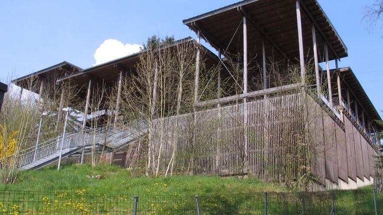 Ein modernes Gebäude auf einem Hang, umgeben von Bäumen und Gras. Die Struktur hat ein Dach mit Überständen und einen Rampenzugang.