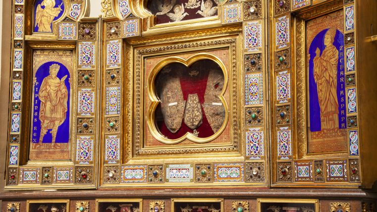 Ein kunstvoll gestaltenes Altarkreuz mit goldenen Verzierungen und bunten Mosaiken. Im Hintergrund sind religiöse Figuren und Symbole sichtbar.